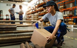 Young woman working in warehouse and taping cardboard box for shipment. Female warehouse worker taping cardboard box with tape dispenser before the shipment.